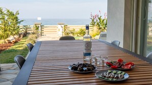 A wooden table with a bottle of liquor, two plates of food, and a glass of water, set against a backdrop of a clear sky and a view of the sea. - Sea and Sun Luxury Pools' Villas (Zagora-Mouresi)