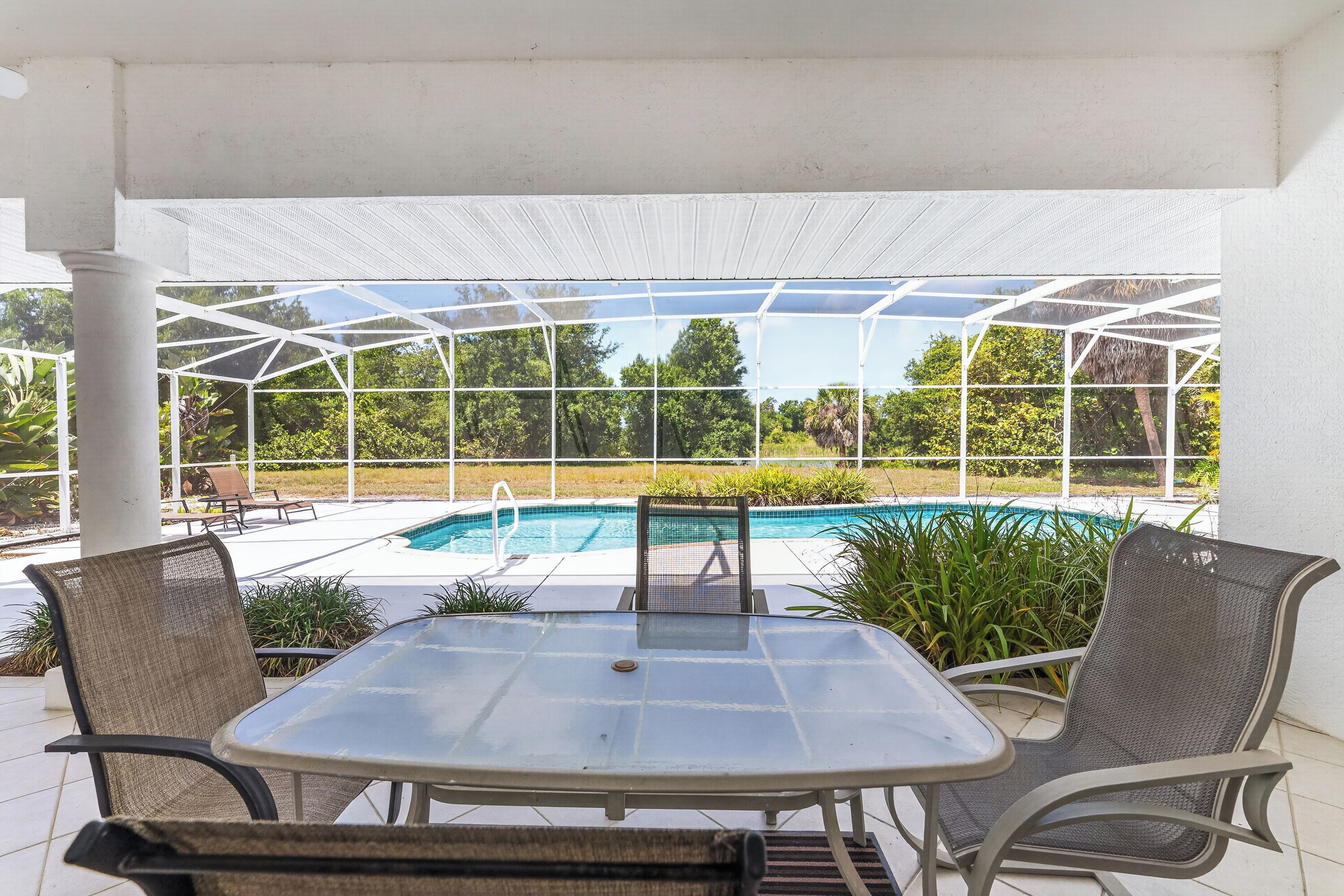 A covered patio area with a glass table and chairs, a pool, and surrounding greenery.