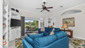 A living room with a blue sofa, a ceiling fan, a flat-screen TV, and a dining area with outdoor seating. - WATERFRONT, HEATED POOL, 3/2/2  single-family Rotonda West Home (Rotonda West)