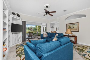 Ein Wohnzimmer mit blauem Sofa, Deckenventilator, Flachbildfernseher und Essbereich mit Sitzgelegenheiten im Freien.