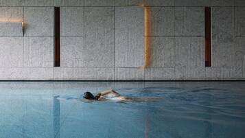 Piscina com uma pessoa nadando, parede texturizada com aberturas retangulares e reflexo na superfície da piscina.