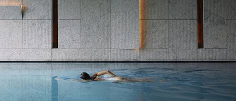 A swimming pool with a person swimming, a textured wall with rectangular openings, and a reflection on the pool's surface.
