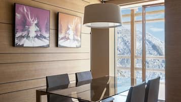 Ein moderner Essbereich mit einem Glastisch, dunklen Stühlen und einer Pendelleuchte. An der Wand hängen zwei gerahmte Kunstwerke und durch große Fenster hat man Blick auf schneebedeckte Berge.
