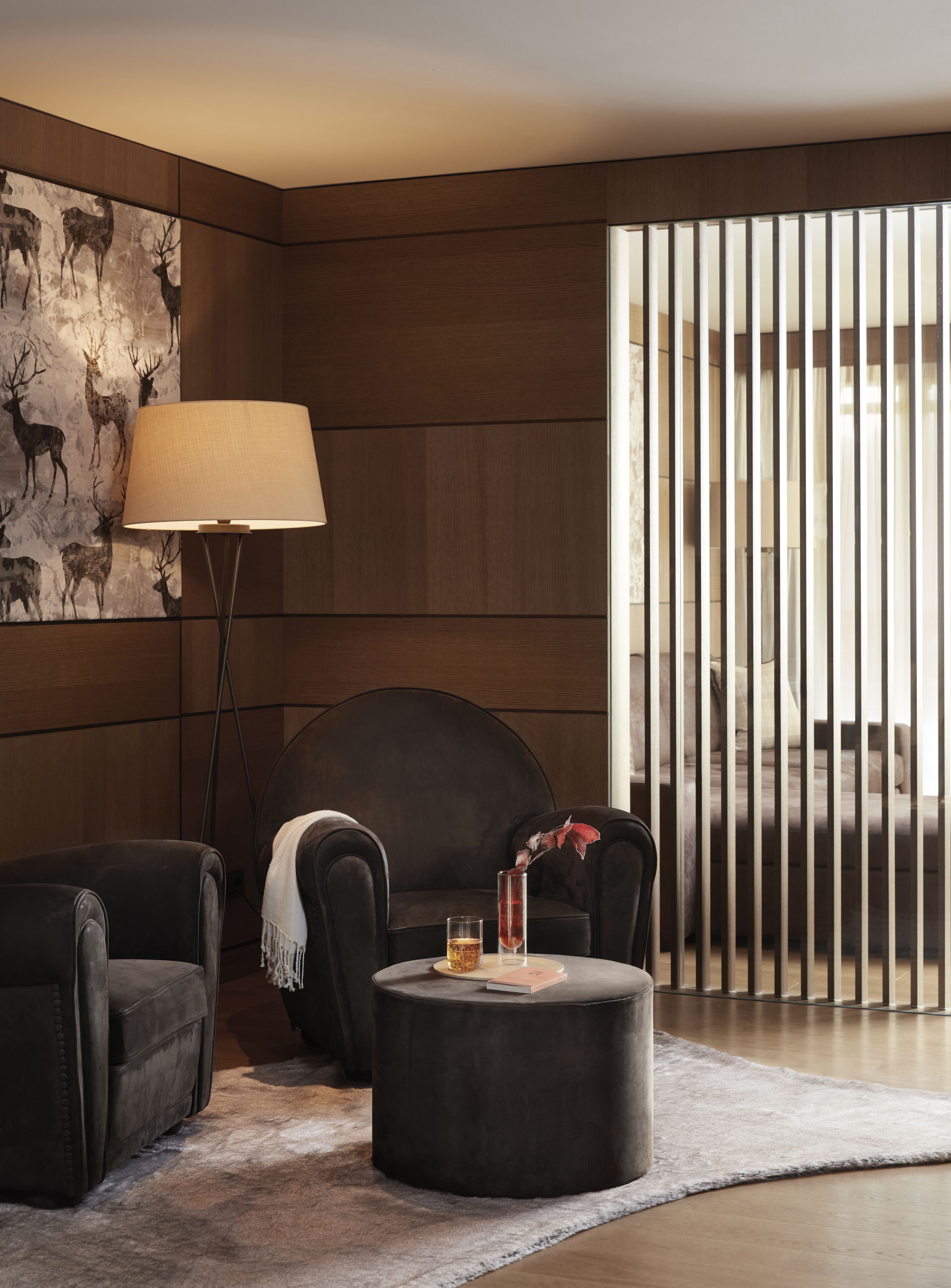 Ein modernes Wohnzimmer mit Wandpaneelen aus dunklem Holz, einer großen Stehlampe, zwei Ledersesseln, einem runden Hocker mit einem Bierglas und einer Vase sowie einem großen Fenster mit vertikalen Rollläden.