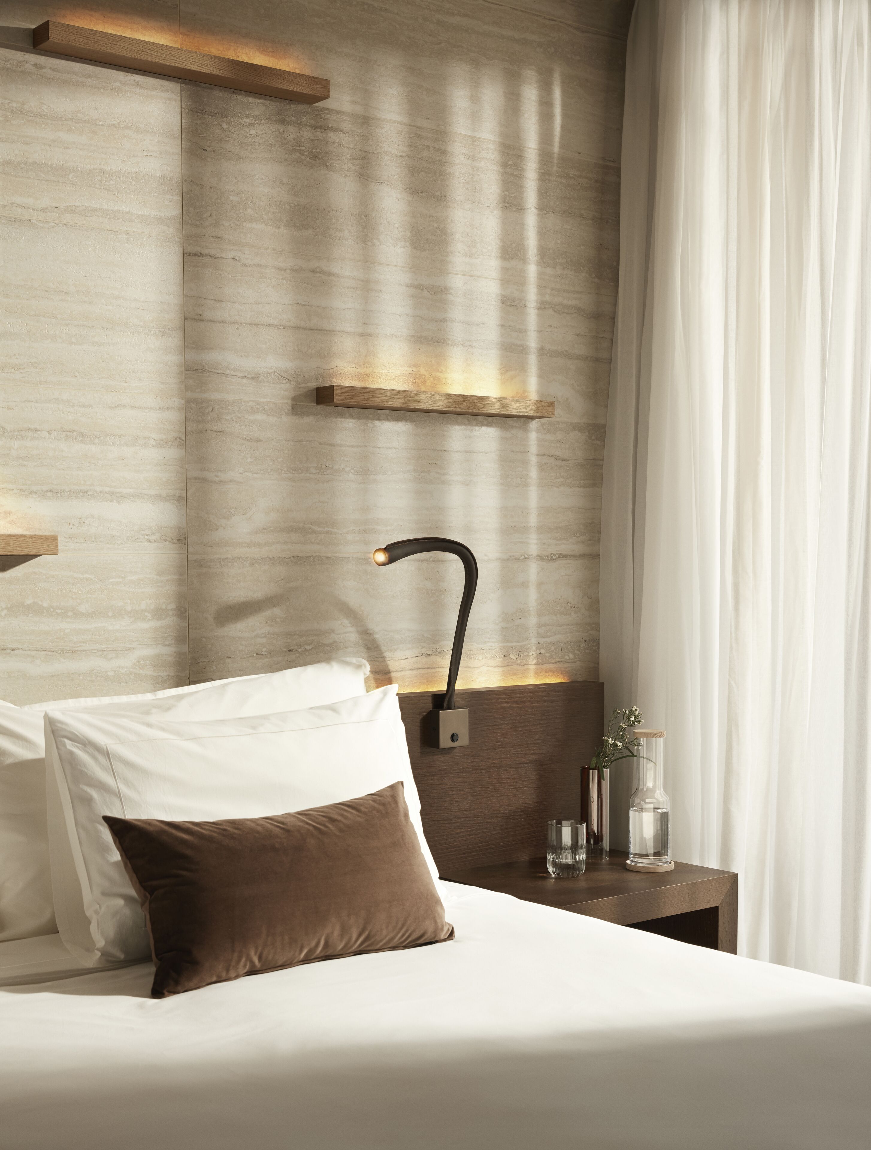 A hotel room with a bed, a nightstand, a wall-mounted lamp, and a window with curtains.