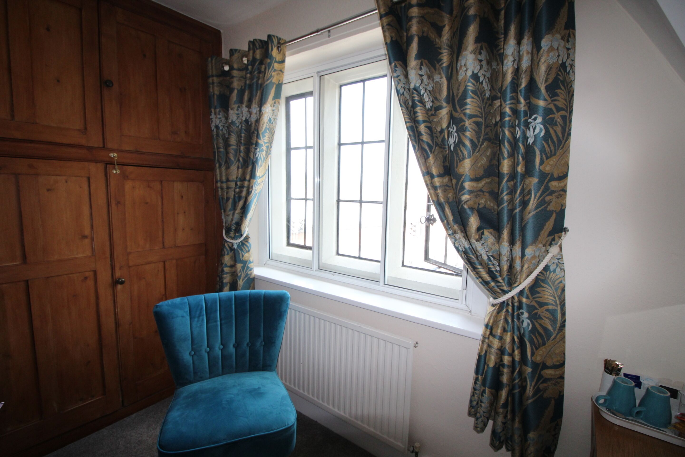 Una habitación con un sillón azul, un armario de madera y una ventana con cortinas florales.