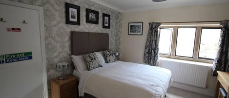 A bedroom with a bed, a nightstand, a lamp, a window with curtains, and a wall with framed pictures.