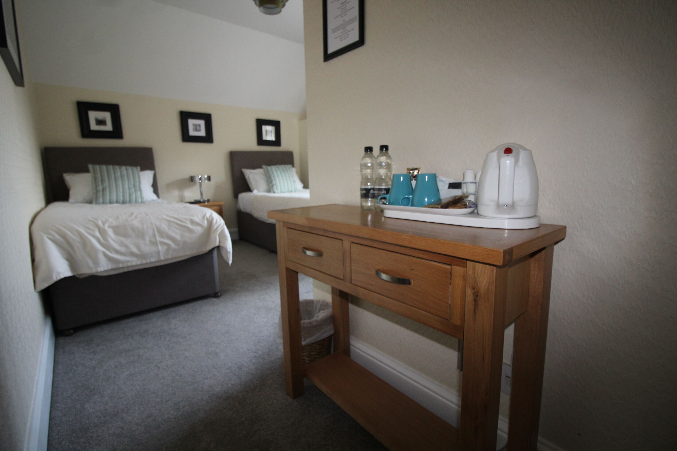 A bedroom with a bed, a wooden nightstand, a desk with a kettle, and two framed pictures on the wall.