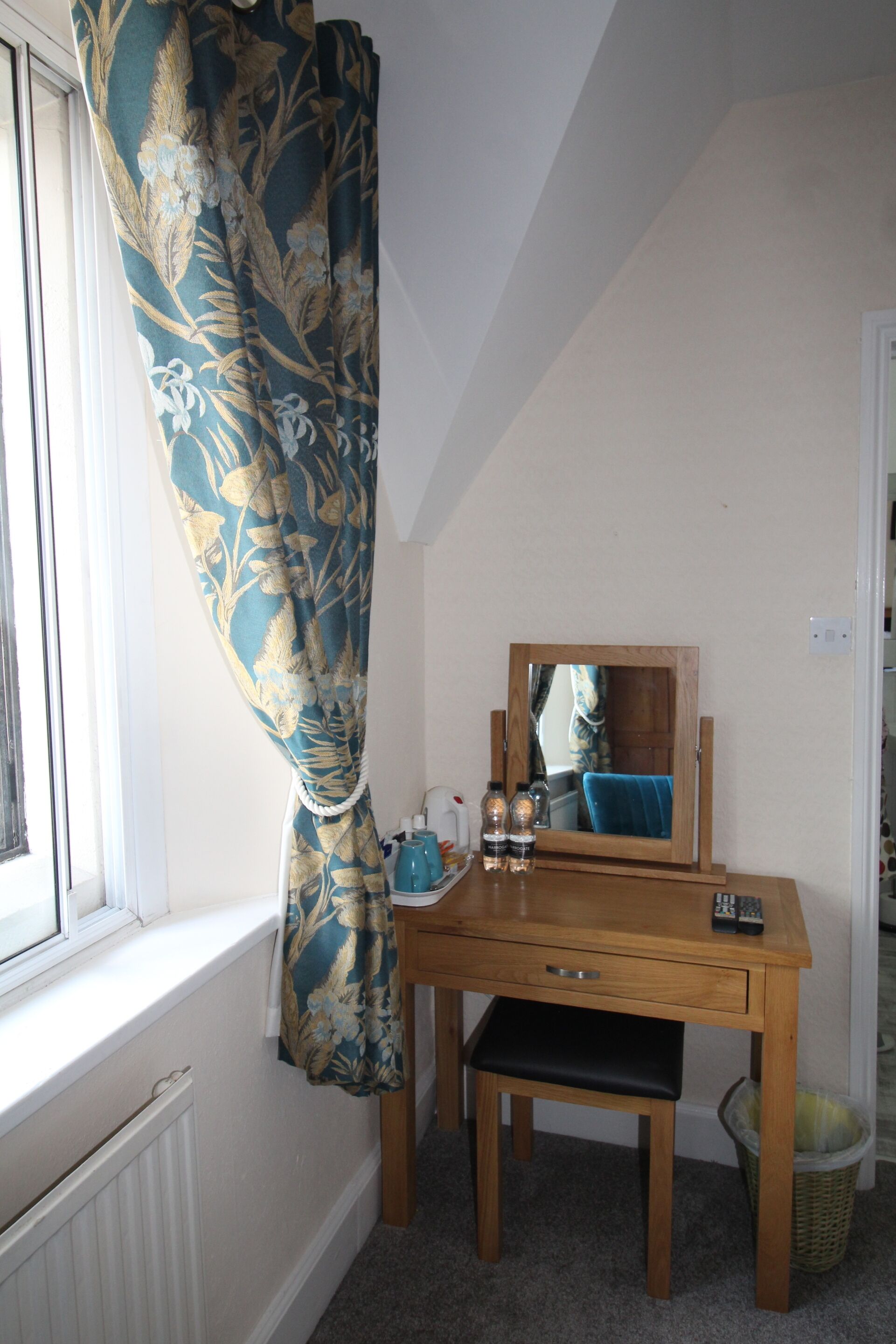 A small room with a wooden desk, a mirror, a window with a curtain, and a radiator.