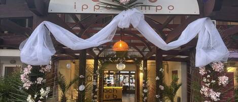 Entrada de hotel con una galería techada, plantas en macetas y un letrero que dice 'bach club'.