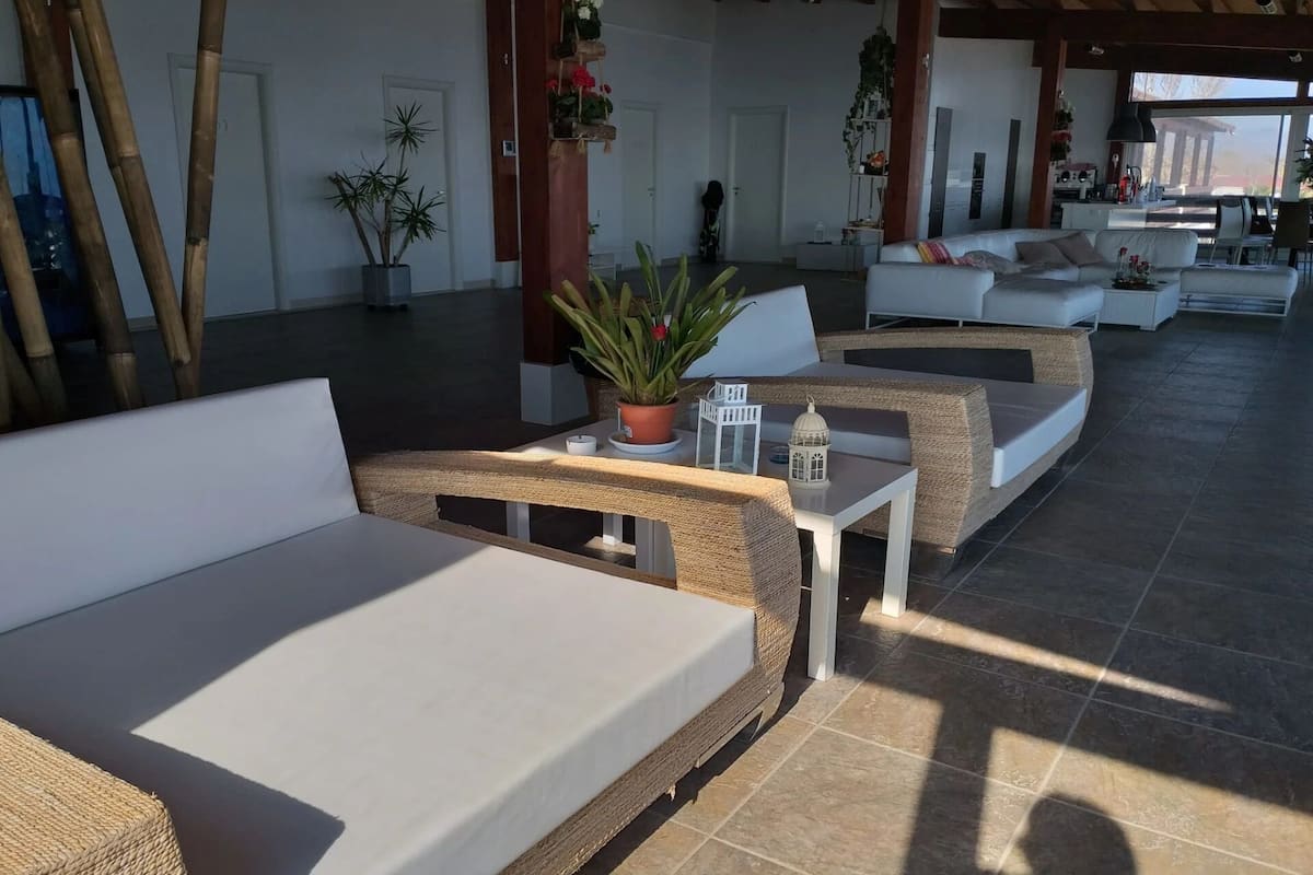 A covered outdoor seating area with wicker furniture and white cushions, surrounded by potted plants and a view of the outside.