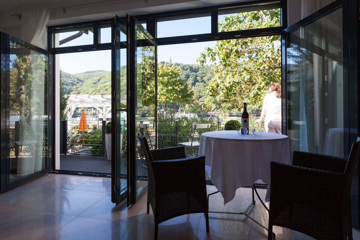 A modern room with large glass doors leading to a balcony, a round table with chairs, and a view of a lush green landscape.