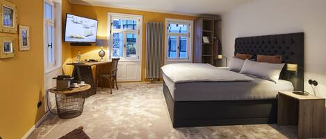Ein Schlafzimmer mit einem großen Bett, einem Schreibtisch mit Stuhl, einem Fernseher und einem Fenster mit Vorhängen.