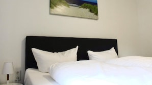 Una habitación de hotel con cama, ropa de cama blanca, una mesita de noche con lámpara y un cuadro de una playa en la pared.