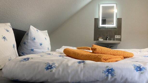A neatly made bed with floral bedding and a pair of orange towels, a bedside table with a mirror, and a wall-mounted sink.