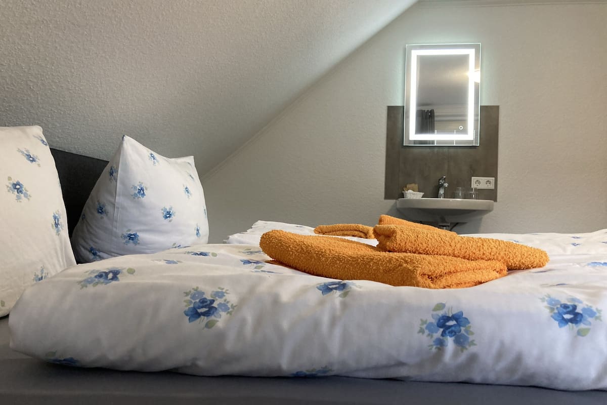 A neatly made bed with floral bedding and a pair of orange towels, a bedside table with a mirror, and a wall-mounted sink.