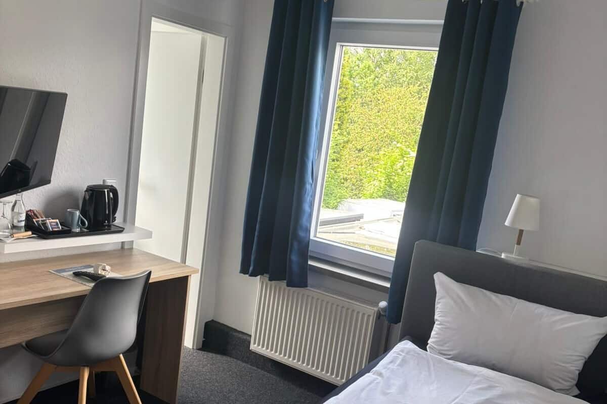 A hotel room with a bed, a desk with a computer, a chair, a window with curtains, and a radiator.
