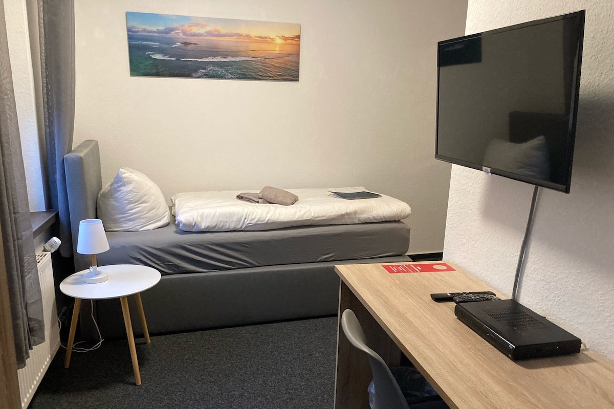 A small room with a bed, a desk, a TV, and a painting on the wall.