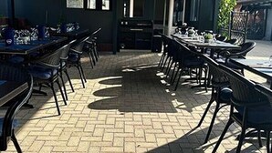 An outdoor dining area with brick flooring and metal chairs. Tables are set with white tablecloths and plates.