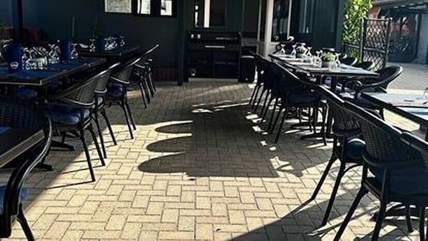 Un área de comedor al aire libre con suelo de ladrillo y sillas de metal. Las mesas están dispuestas con manteles blancos y platos.