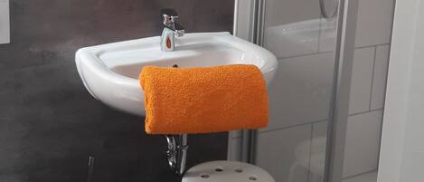 A bathroom with a white sink, a glass shower enclosure, a white toilet, and a small stool with a towel.