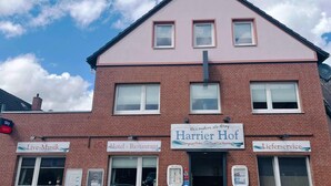 Un edificio de dos plantas con fachada rosa y techo negro. Tiene varias ventanas, algunas con persianas, y carteles que dicen 'Live-Musik', 'Hotel - Restaurant', 'Harrier Hof' y 'Leiferservice'.