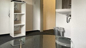 Una habitación moderna con una mesa de vidrio, un estante blanco, un televisor de pantalla plana montado en la pared y un baño con lavamanos y espejo.