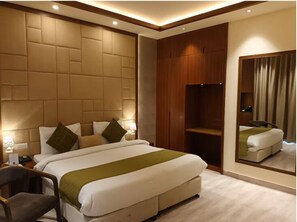 A modern hotel room with a large bed, a wooden wardrobe, a mirror, and a small desk with a chair.