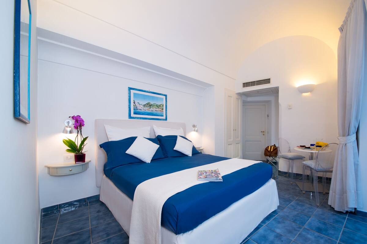A hotel room with a bed, a small table, chairs, a picture on the wall, and a door leading to another room.