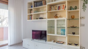 A modern living room with a flat-screen TV on a white cabinet, shelves with books and decorative items, and a glass cabinet with glassware.
