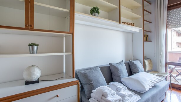 A modern bedroom with a grey sofa, white towels, a small potted plant, and a shelf with a plant and a decorative item.