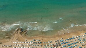 An aerial view of a beach with rows of beach umbrellas and lounge chairs, a rocky shoreline, and clear turquoise water. - Nami Hotel & Suites (Hersonissos)