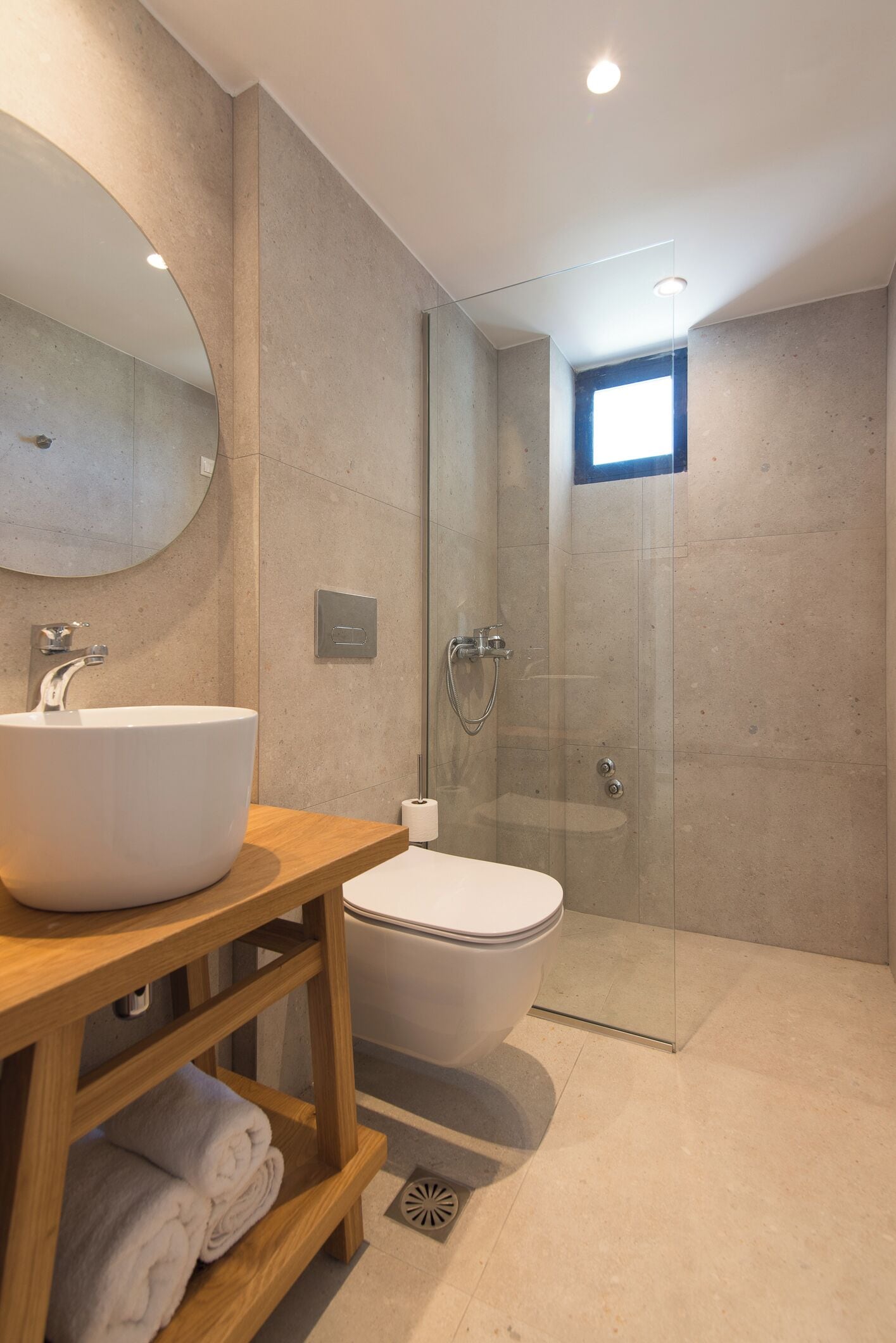 Un baño moderno con un espejo redondo, un tocador de madera, un lavabo blanco, un inodoro y una ducha con mampara de vidrio.