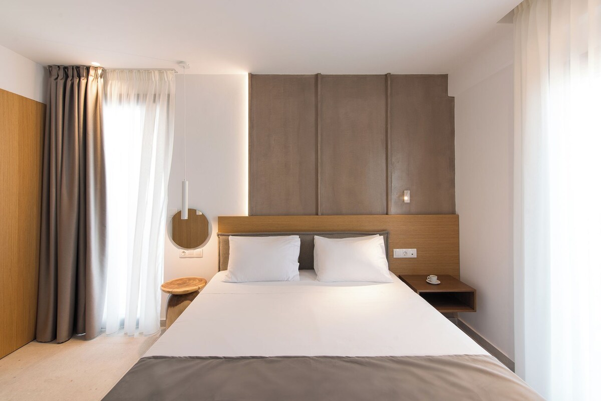 A modern hotel room with a large bed, wooden headboard, bedside tables, and a view of the outdoors.