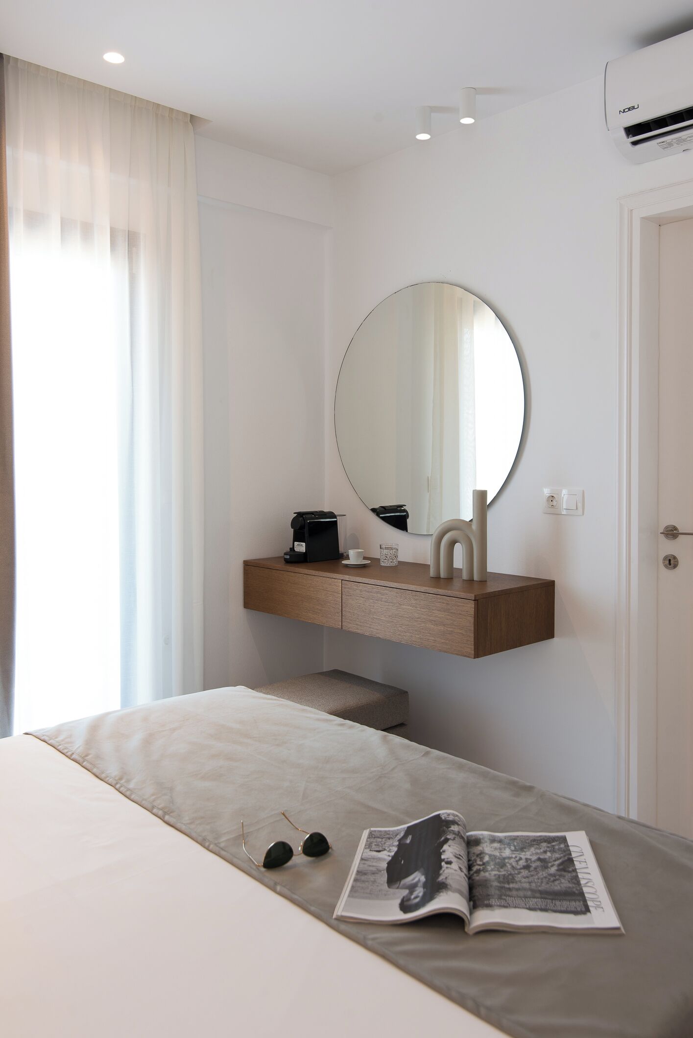 A hotel room with a bed, a bedside table with a coffee maker, a round mirror, and a window with curtains.