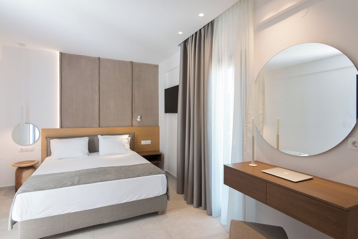 A modern hotel room with a bed, bedside table, mirror, and a large window with curtains.
