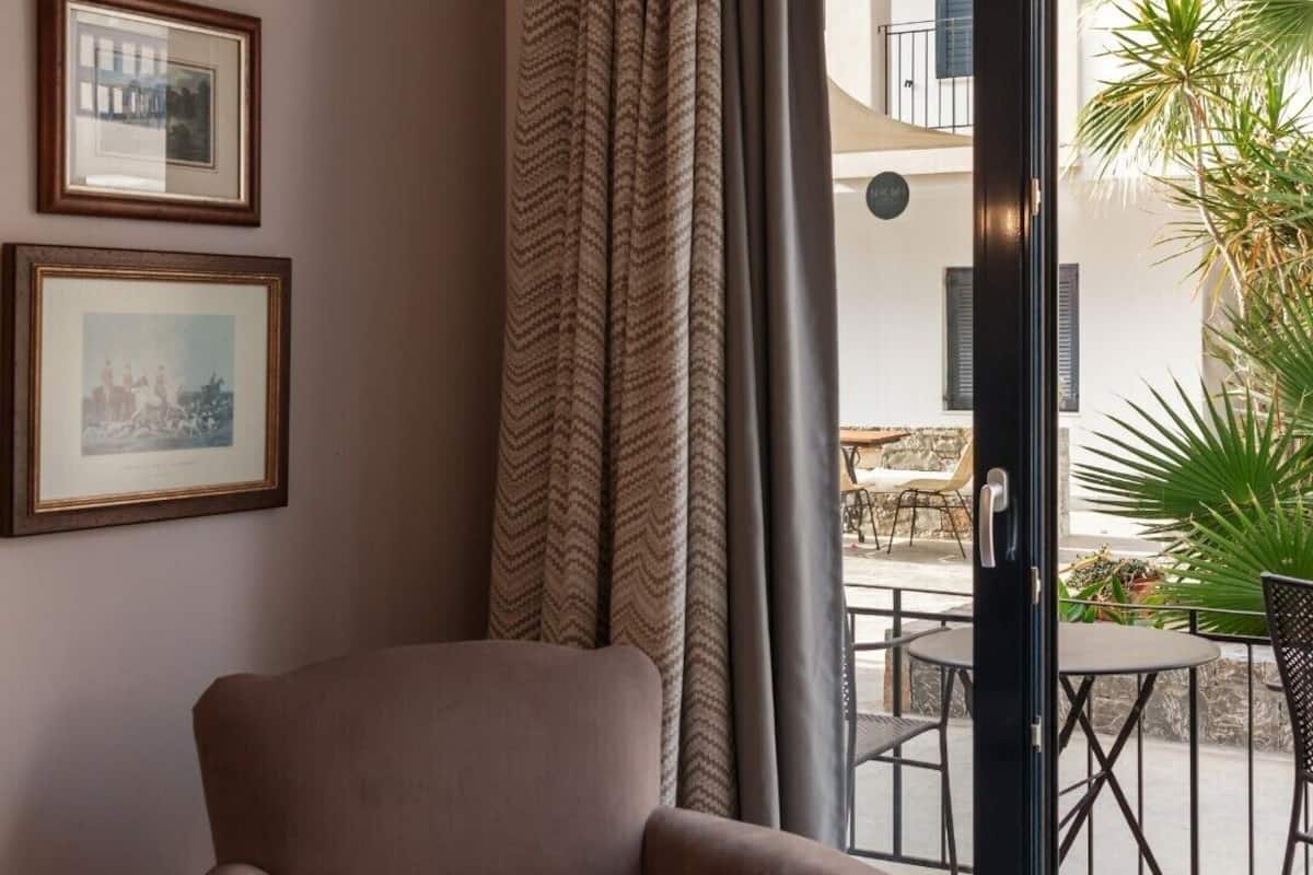 Una habitación con un sillón marrón, un ventanal con cortinas y dos cuadros en la pared.
