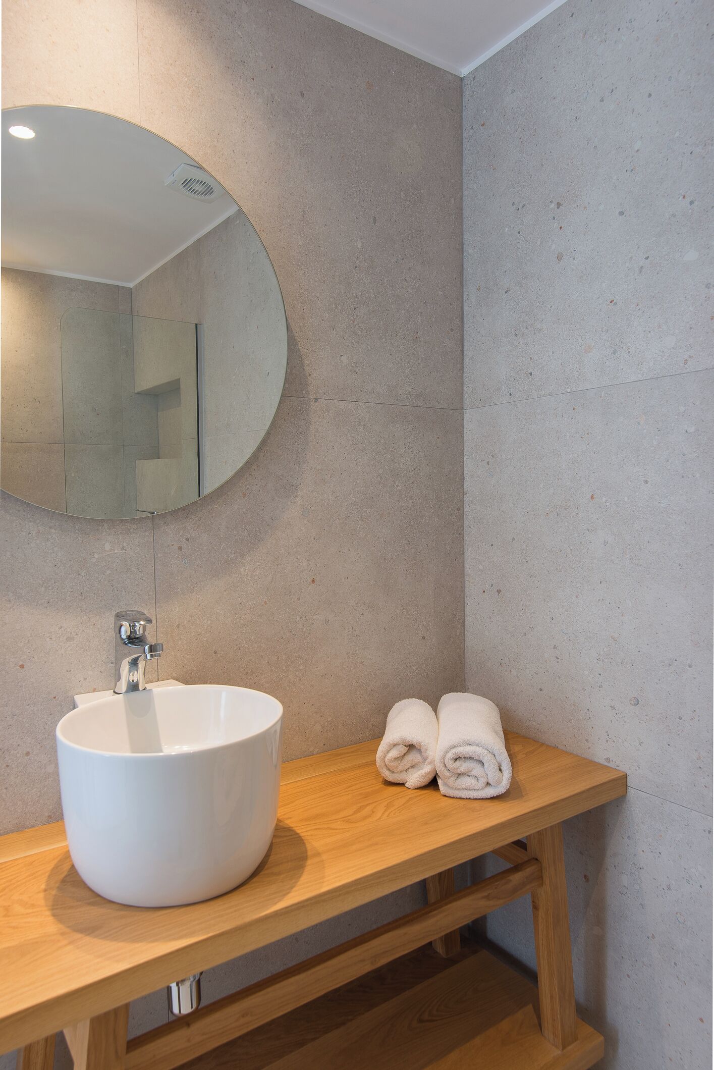 A bathroom with a round mirror, a white sink, and two rolled towels on a wooden vanity.