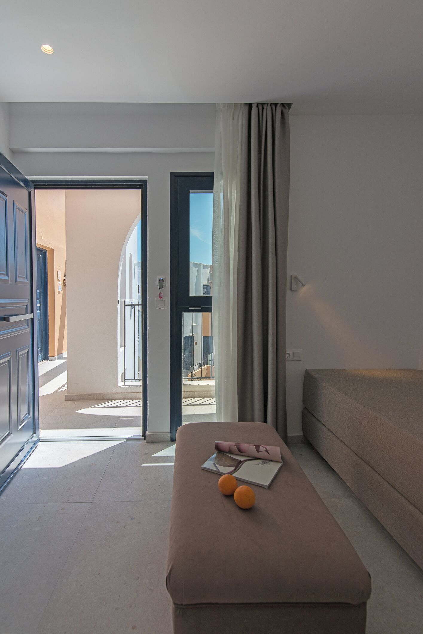 A modern hotel room with a sofa, a small table with books and oranges, a balcony door, and a view of the outside.