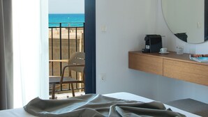 A hotel room with a bed, a desk with a coffee maker, a chair, a large mirror, and a view of the ocean through an open door. - Nami Hotel & Suites (Hersonissos)