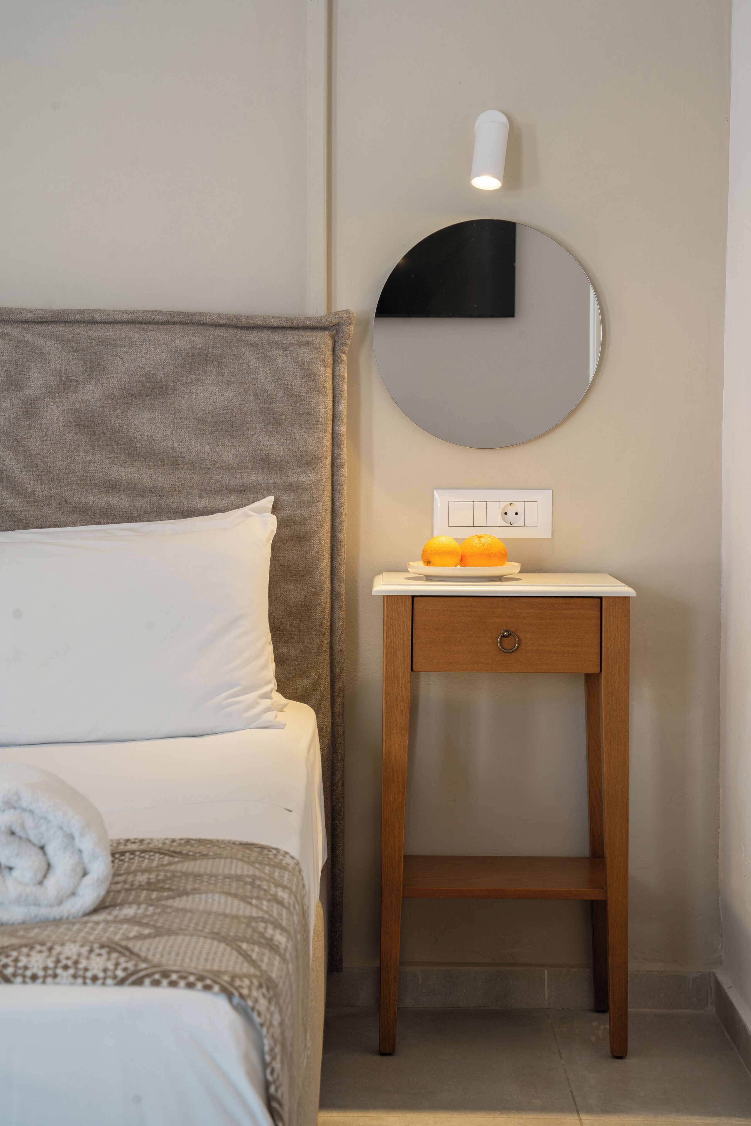 Een netjes opgemaakt bed met een kussen en een opgerolde handdoek, een nachtkastje met een bord mandarijnen, een wandspiegel en een lamp.