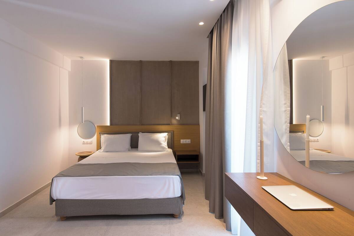 Una habitación de hotel moderna con una cama grande, mesitas de noche, un espejo y un ordenador portátil sobre el escritorio.