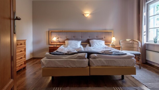 Una cama doble con dos mesitas de noche, una lámpara de pared, un sillón de mimbre y una ventana con cortinas.