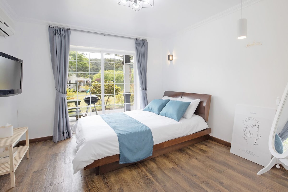 A bedroom with a bed, a TV, a window with curtains, a chair, and a fan.