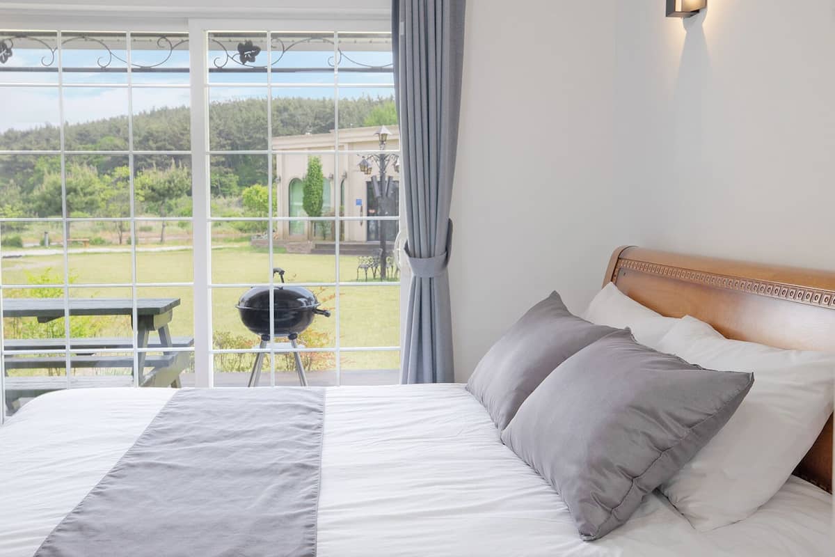 A bedroom with a large window, a bed with grey bedding, and a view of a garden and building outside.
