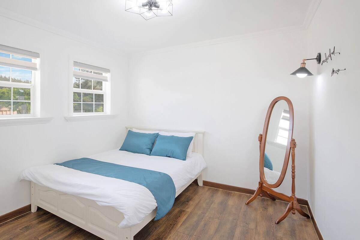 A bedroom with a bed, two windows, a ceiling fan, a mirror, and a wooden floor.