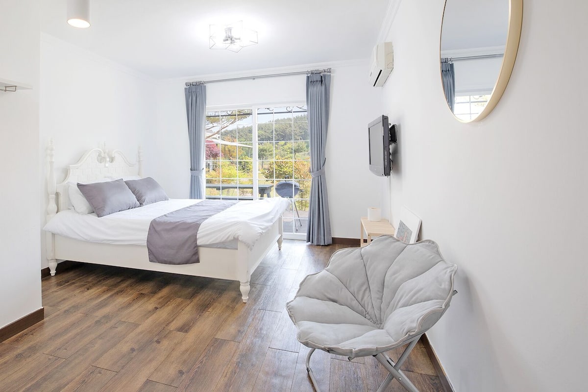 A bedroom with a bed, a chair, a TV, and a window with curtains.