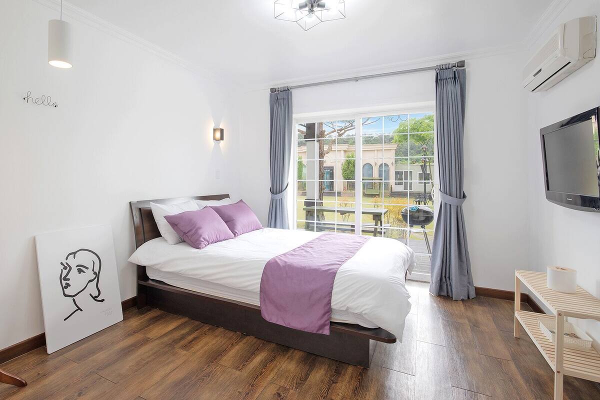 A bedroom with a bed, a painting, a wooden floor, a window with curtains, and a TV.