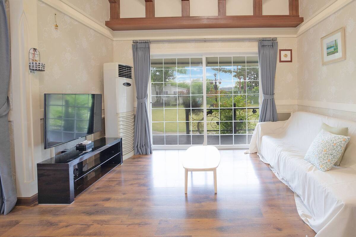 A living room with a TV, a white sofa, a small table, and a large window with curtains.