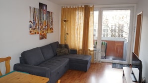 Een woonkamer met een grijze modulaire bank, een houten salontafel en een radiator. Er hangen drie ingekaderde foto's aan de muur, een raam met gordijnen en een deur naar buiten.